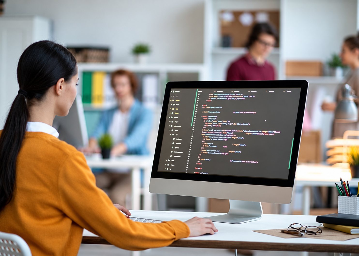 Une jeune femme aux cheveux attachés, vue de dos, travaille sur un ordinateur affichant du code dans un bureau lumineux où des collègues discutent en arrière-plan.
