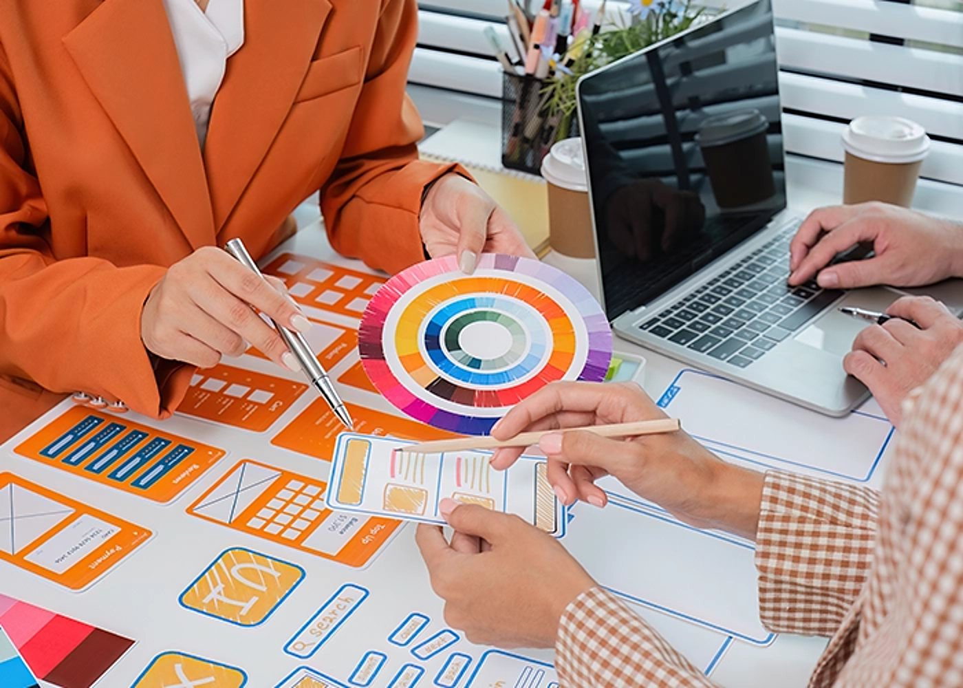 Autour d'une table couverte de maquettes d'interface orange et bleues, des personnes tiennent un nuancier circulaire et travaillent sur un ordinateur portable.