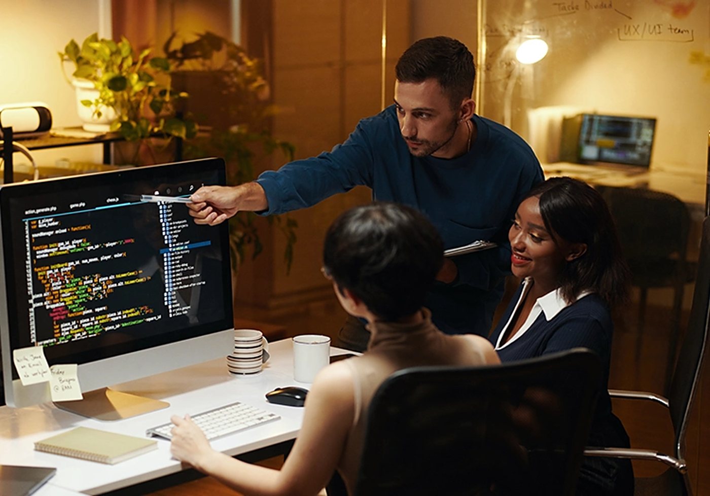 Trois collègues réunis autour d'un bureau regardent du code affiché sur un écran d'ordinateur, dans un bureau chaleureux en soirée.