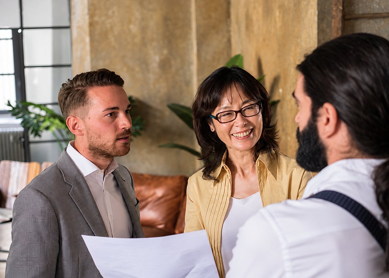 Un homme en veste, une femme souriante à lunettes et un homme barbu de dos regardent une feuille de papier dans un espace aux murs patinés et aux plantes vertes.